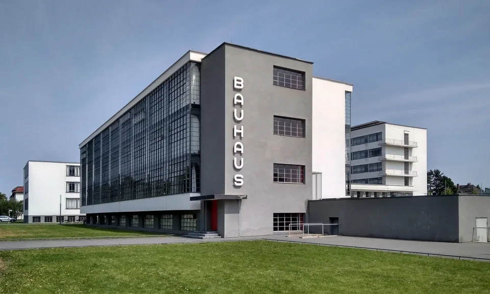 A modernist building with a continuous glass curtain wall and the vertical lettering BAUHAUS, with a lawn in front