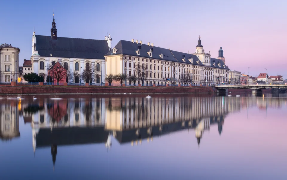 A long university facade and a church with a tall roof on the riverbank, the calm river reflecting the buildings at dusk