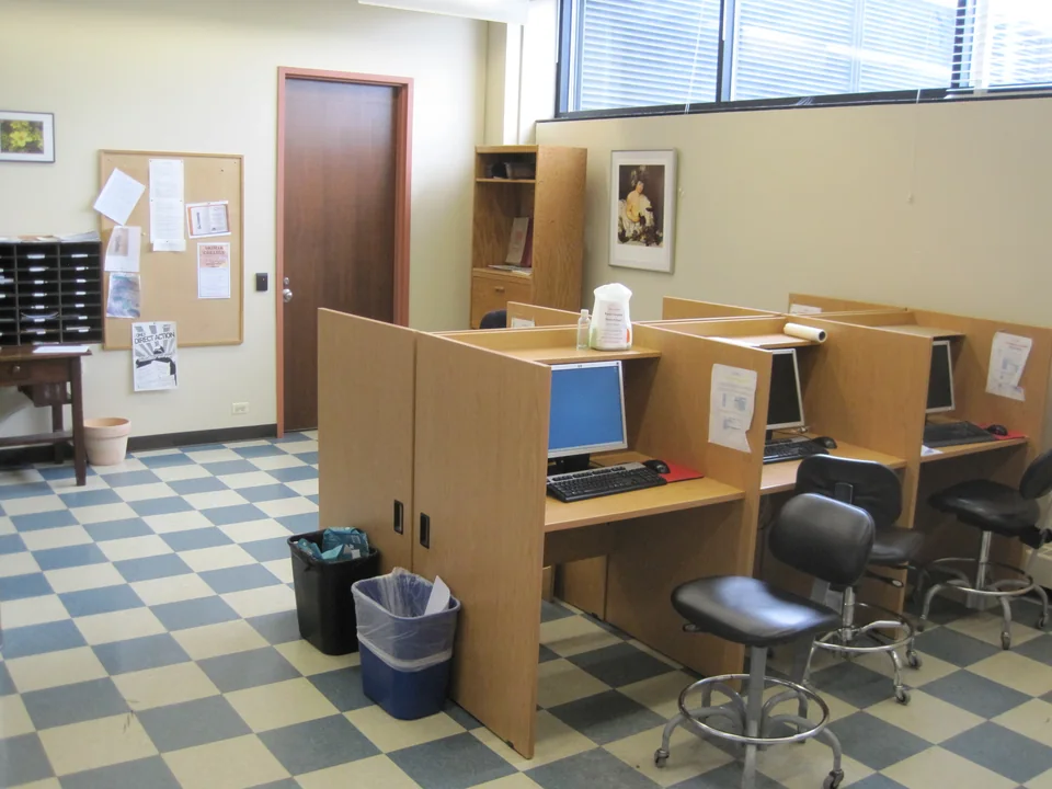 Wooden carrels with monitors, keyboards and chairs on a checkered floor, with a noticeboard on the wall
