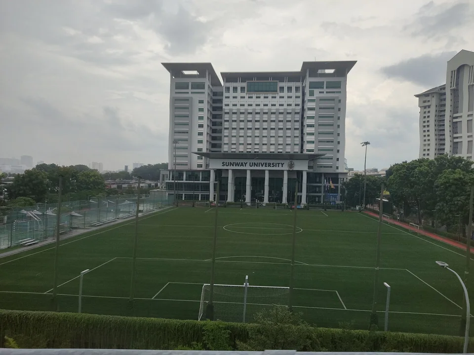Многоэтажный главный корпус Sunway University за пустым футбольным полем кампуса в пасмурный день