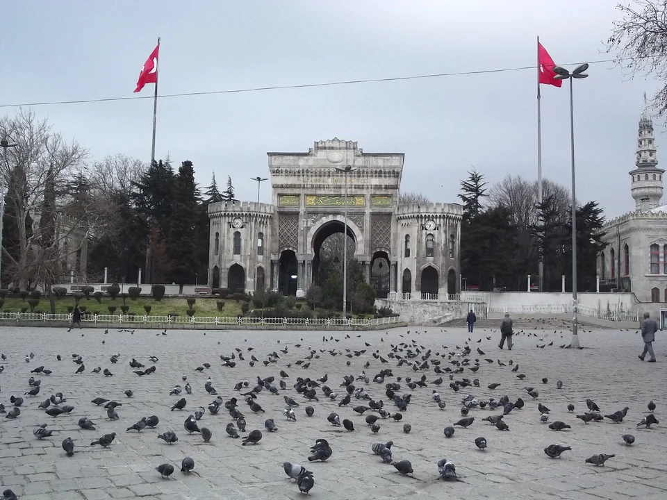 Мавританские ворота с часовыми башенками и надписью İstanbul Üniversitesi, по бокам флаги Турции, на площади голуби