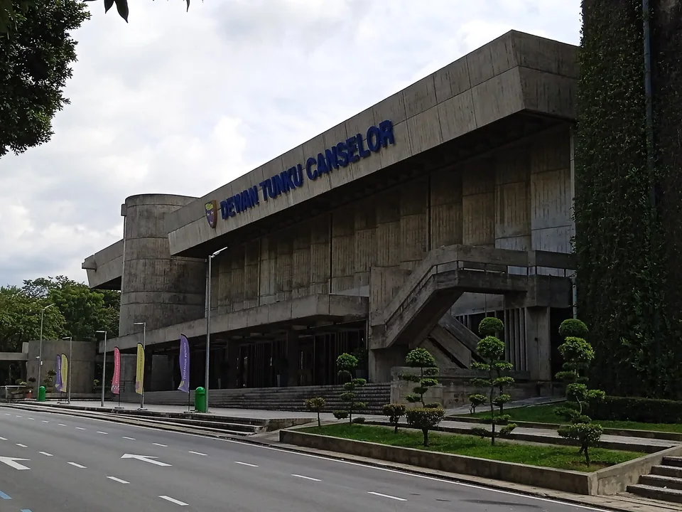 Бетонное здание зала Dewan Tunku Canselor Университета Малайя с лестницей, флагами и подстриженными деревьями у дороги