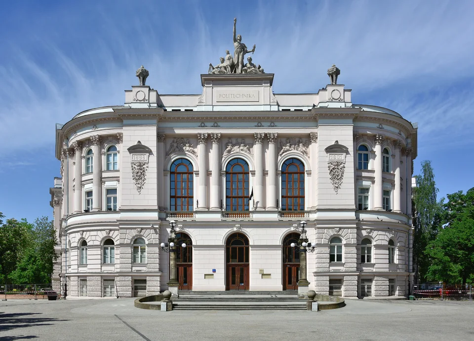 A pale building with rounded corners, arched windows and a sculpture group on the roof, with an empty square in front