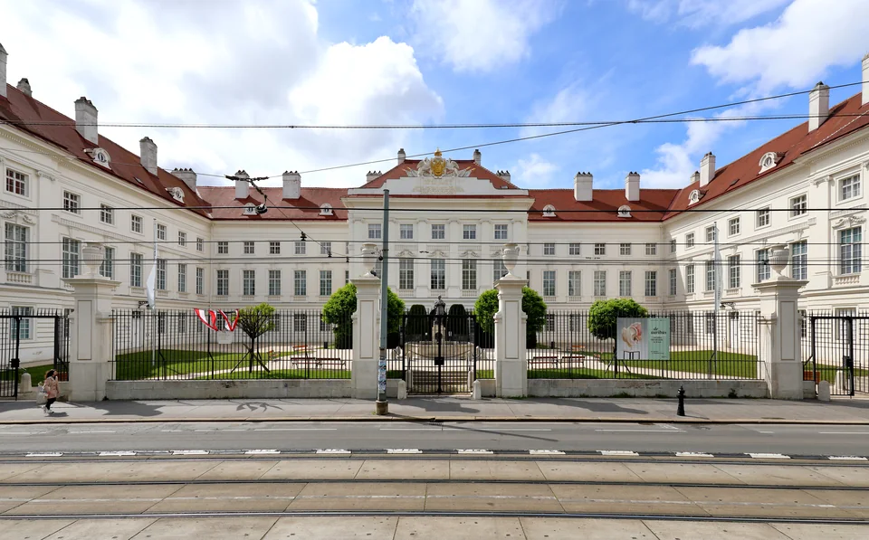 Белое барочное здание с красной крышей и центральным ризалитом за кованой оградой, на улице трамвайные пути