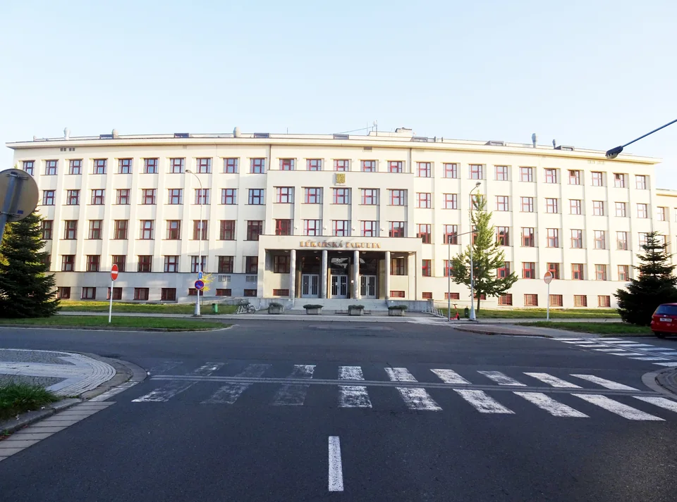 Длинное светлое здание с рядами окон и надписью Lékařská fakulta над входом, перед ним перекрёсток с пешеходным переходом