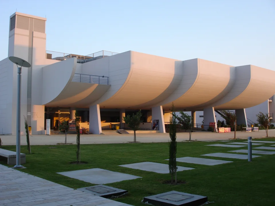 A white building with a curved overhanging upper section on columns, with a lawn crossed by concrete stepping slabs and young trees in front