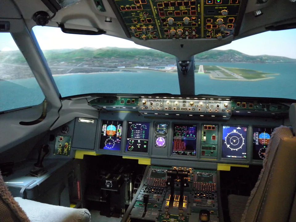 Airliner cockpit with digital displays and an overhead panel, a simulated approach to a seaside runway in the windows