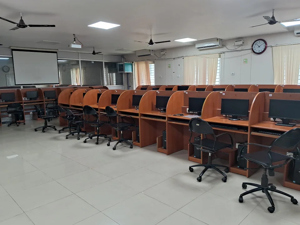 Rows of wooden carrel desks with monitors and black office chairs, a projector screen on the wall