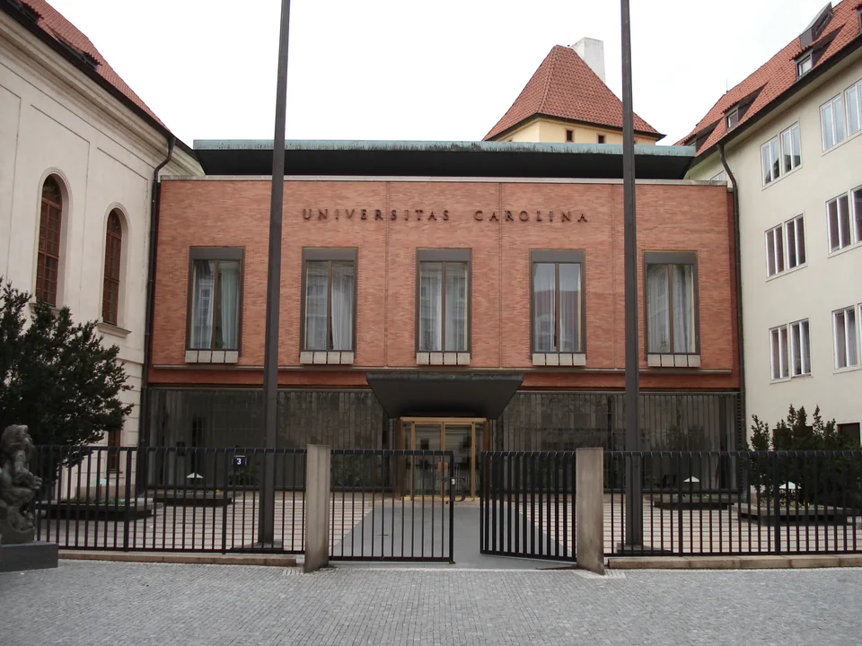 Кирпичный фасад с надписью Universitas Carolina между светлыми боковыми корпусами, перед ним металлическая ограда и флагштоки