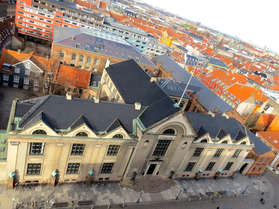 A view from above of a building with a dark roof and a pale façade, with the red-tiled roofs of the city behind it