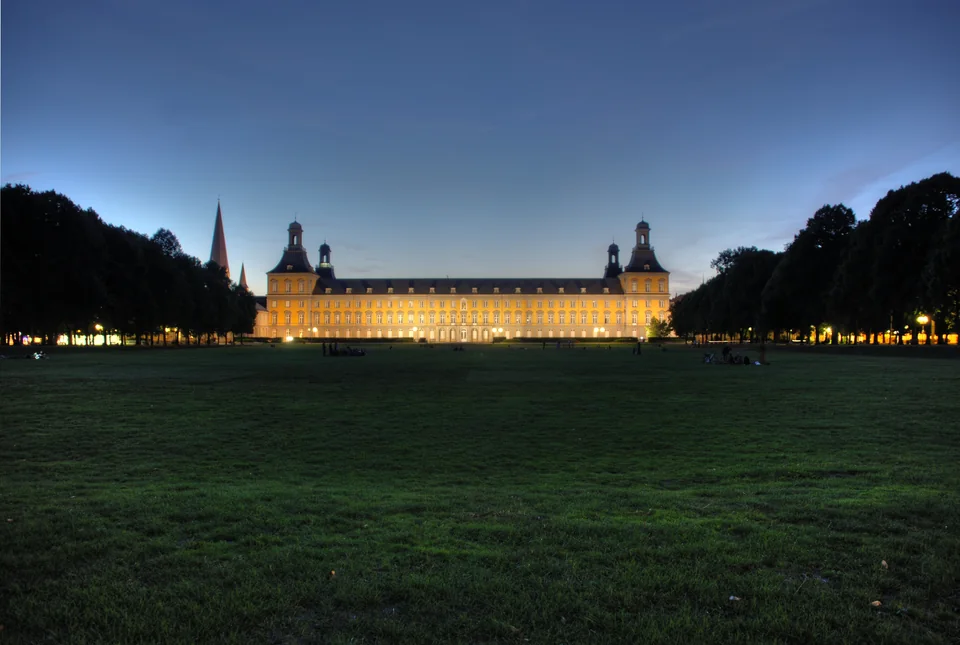 A long illuminated palace with two towers at the edge of a large dark lawn, with trees on both sides and an evening sky
