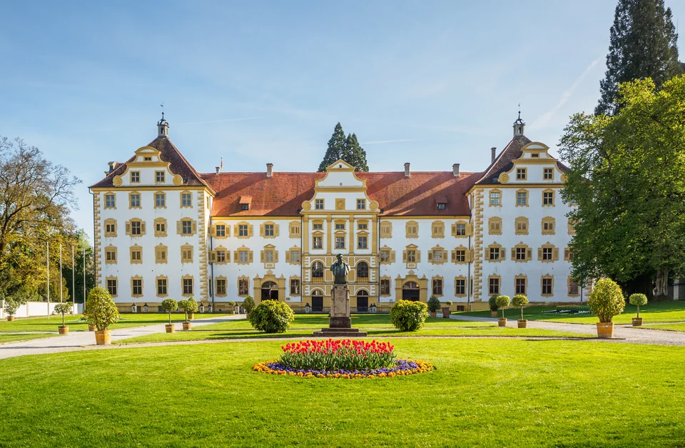 A Baroque palace with a white and ochre façade and turrets, with a lawn, a statue and a tulip bed in front