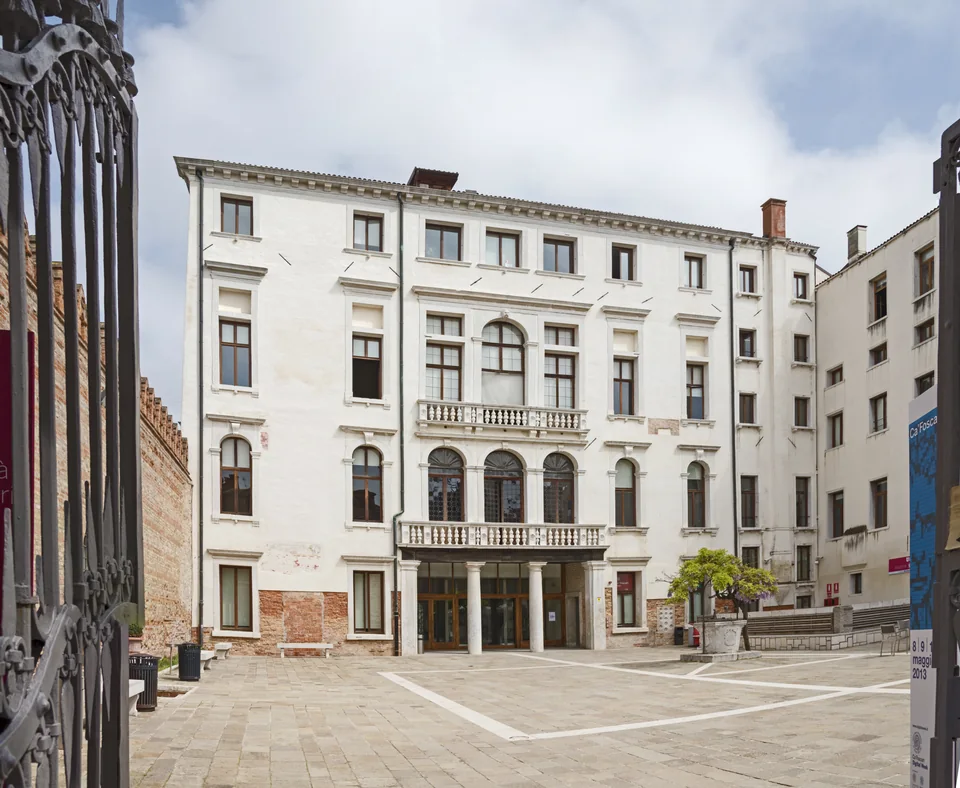 A white palazzo with balconies and arched windows, an empty paved courtyard in front and a wrought-iron gate on the left