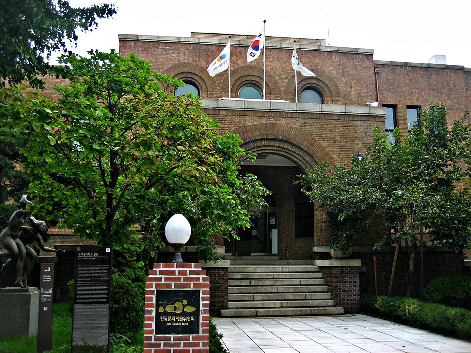 Brick building with an arched entrance and steps behind trees, signboards and a lamp at the entrance