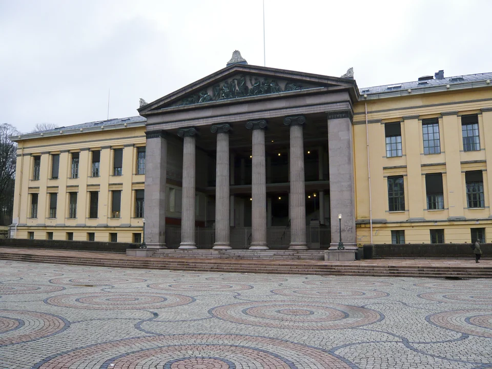 A yellow building with a granite portico of Ionic columns and a sculpted pediment, with a square of patterned paving in front