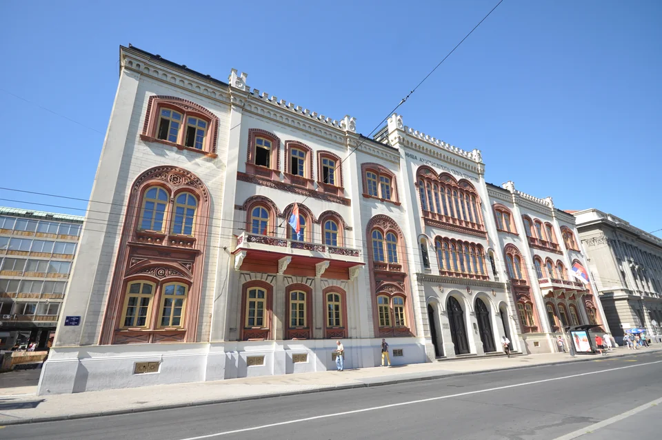 An ornate building with a red-and-cream façade, arched windows and flags, with a street and a few distant passers-by alongside
