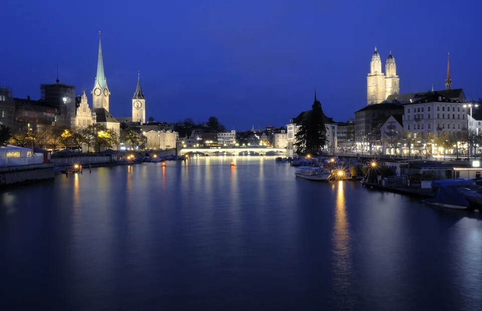 A river in blue twilight lined with lit buildings, a church spire and a twin-towered minster