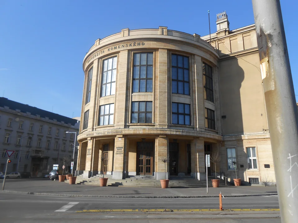A curved stone building with the words Univerzita Komenského, tall windows, wide steps and potted trees, with an empty street in front