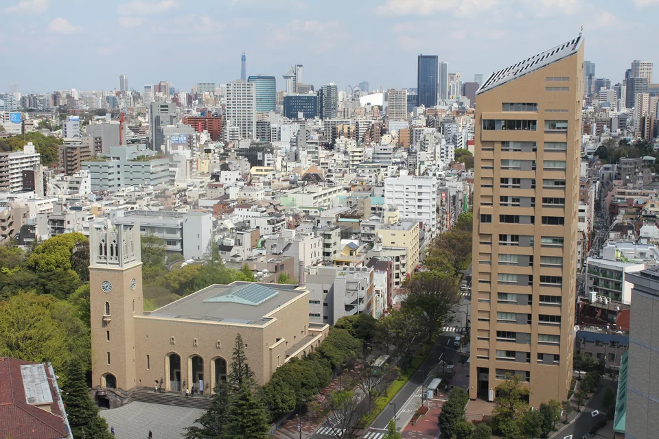 A view from above of a building with a clock tower and a tall beige block, surrounded by dense Tokyo city blocks