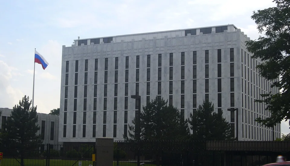 Многоэтажное здание из белого мрамора с узкими вертикальными окнами за оградой, слева флаг России на флагштоке