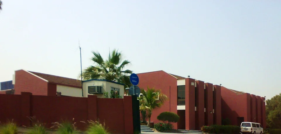 Кирпично-красные корпуса Dubai College за оградой у главного въезда, пальмы и знак Entry