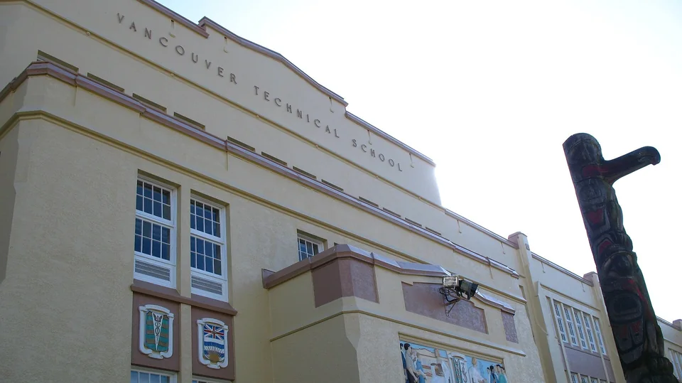 Светлый фасад школы в стиле ар-деко с надписью Vancouver Technical School, гербами и росписью, справа резной тотемный столб