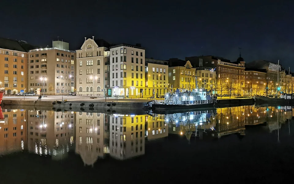 A row of lit buildings along a waterfront and a moored ship, their lights reflected in still water