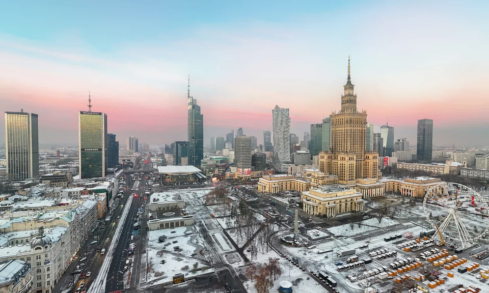 An elevated view of a snow-covered city with wide streets, high-rise towers and the spire of the Palace of Culture against a pink sky