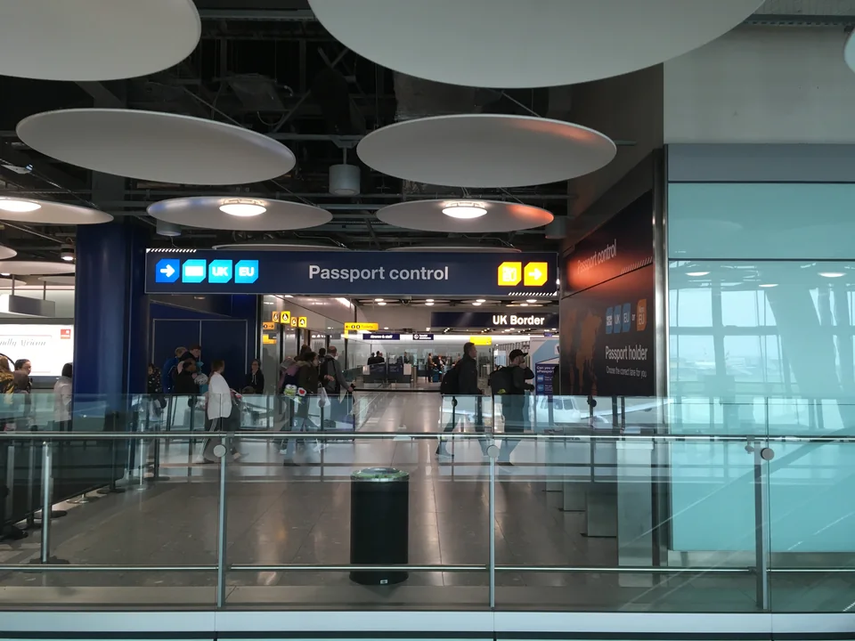 Светлый зал аэропорта с указателями Passport control и UK Border, стеклянными ограждениями и путешественниками