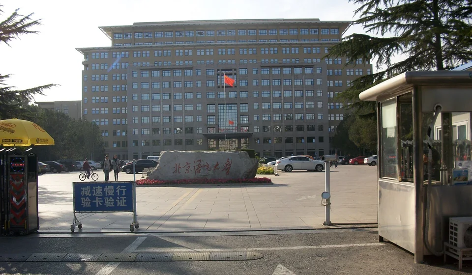Вид через въездные ворота на камень с надписью «北京语言大学» и многоэтажный главный корпус с красным флагом, слева синий указатель, справа будка охраны