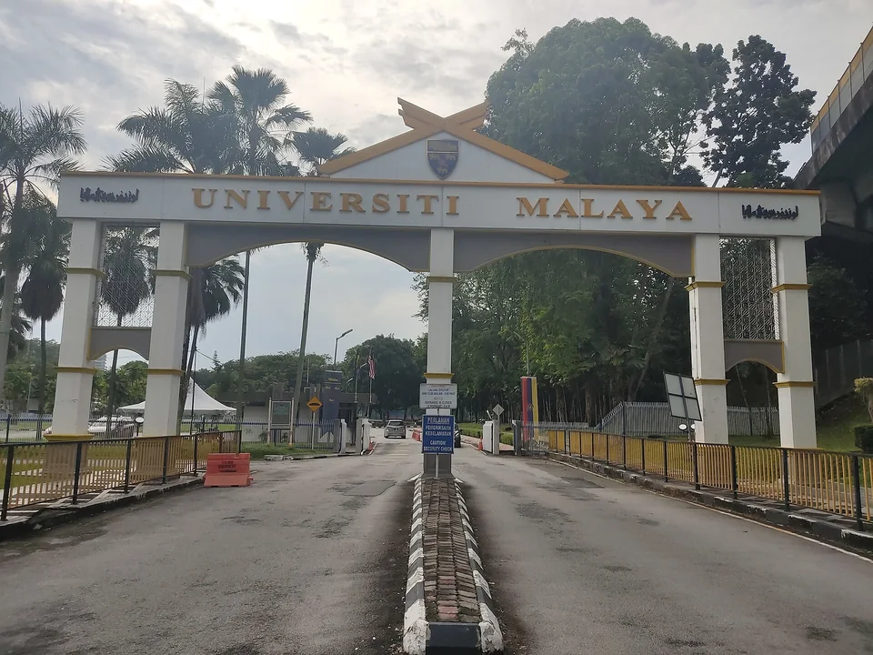 Белая арка ворот с надписью Universiti Malaya и гербом, за ней въезд в кампус среди пальм и тропических деревьев