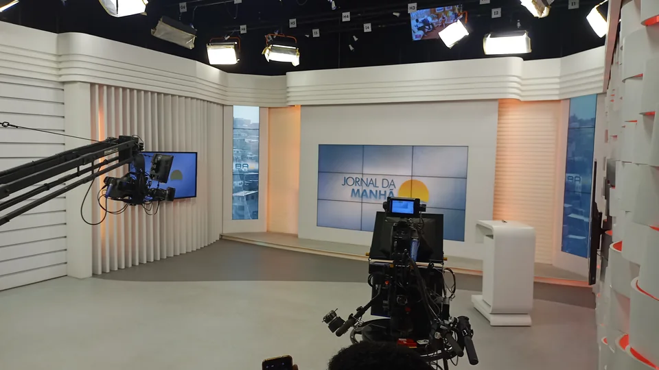 An empty bright TV studio with a screen and presenter's desk, a studio camera and crane in the foreground and rows of lights above