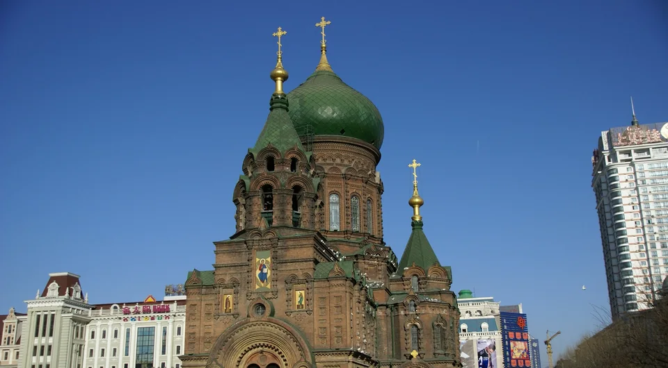 Кирпичный собор с зелёным куполом и золотыми крестами на фоне ясного синего неба, рядом здания центра Харбина