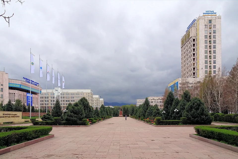 Широкая мощёная аллея между елями и подстриженными кустами, слева флагштоки и корпуса, справа высотное здание университета