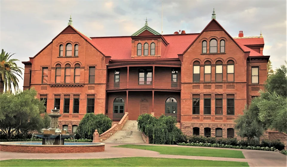 A three-storey red-brick building with gables and a veranda, with a fountain, lawn and palm trees in front