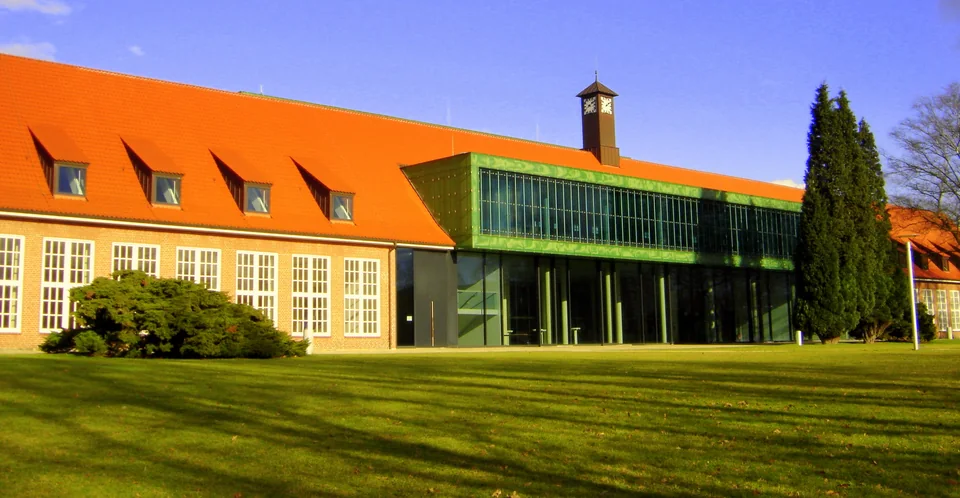 A brick building with an orange tiled roof and a glazed extension, with a sunlit lawn in front