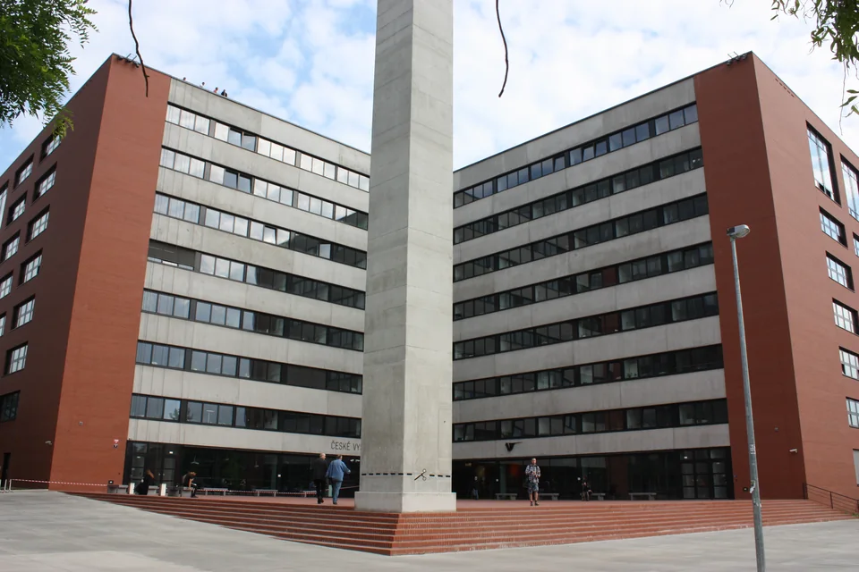 Два крыла современного корпуса ČVUT с красно-коричневой облицовкой и ленточными окнами, между ними высокий бетонный обелиск и широкая лестница