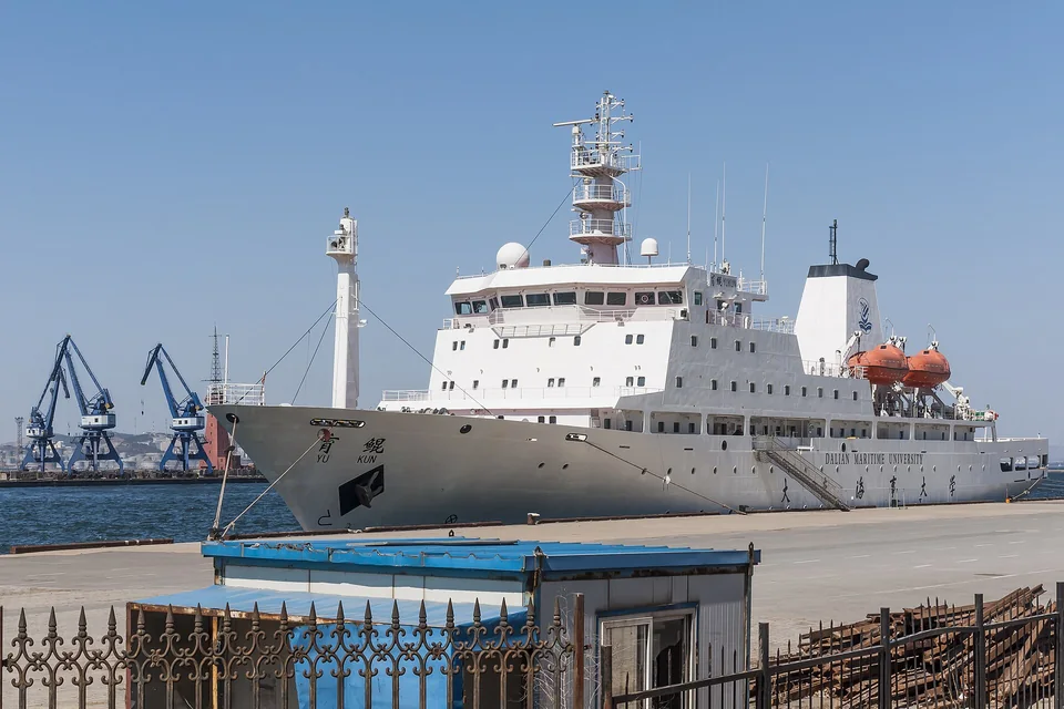 Белое учебное судно с надписью Dalian Maritime University на борту стоит у причала, слева портовые краны