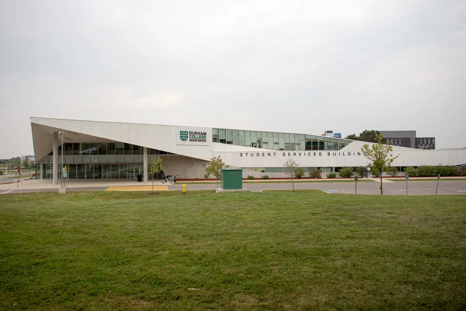 Белое здание с наклонной крышей, стеклянным фасадом, логотипом Durham College и надписью Student Services Building, перед ним большой газон