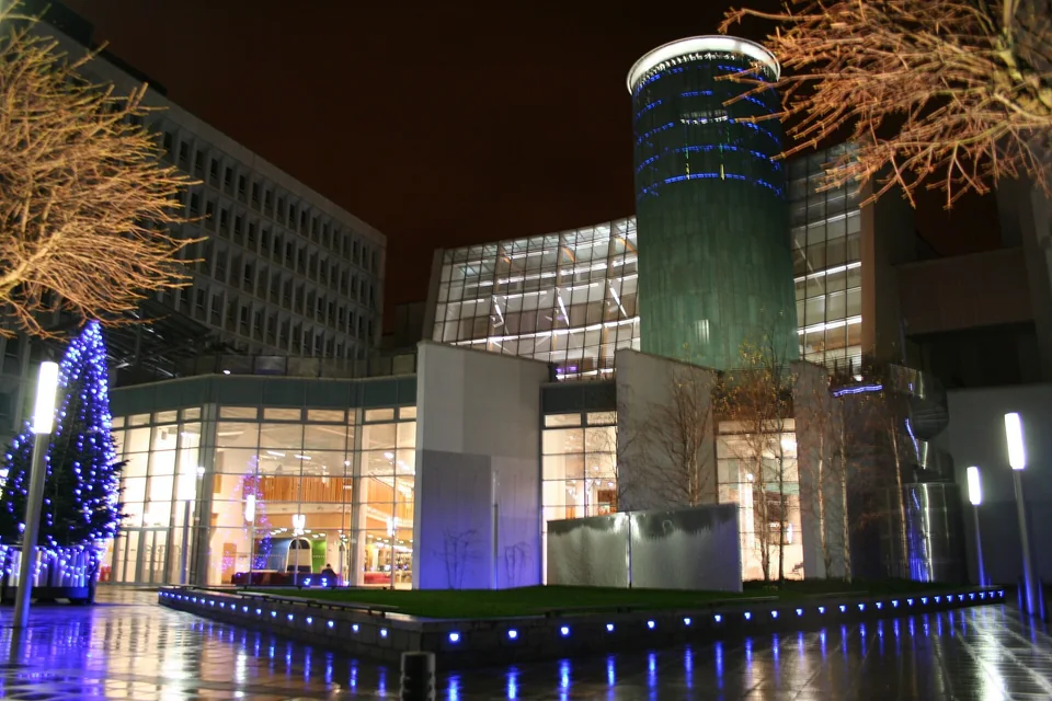 Освещённое ночью стеклянное здание Saltire Centre с цилиндрической башней и подсвеченной ёлкой на площади кампуса GCU в Глазго