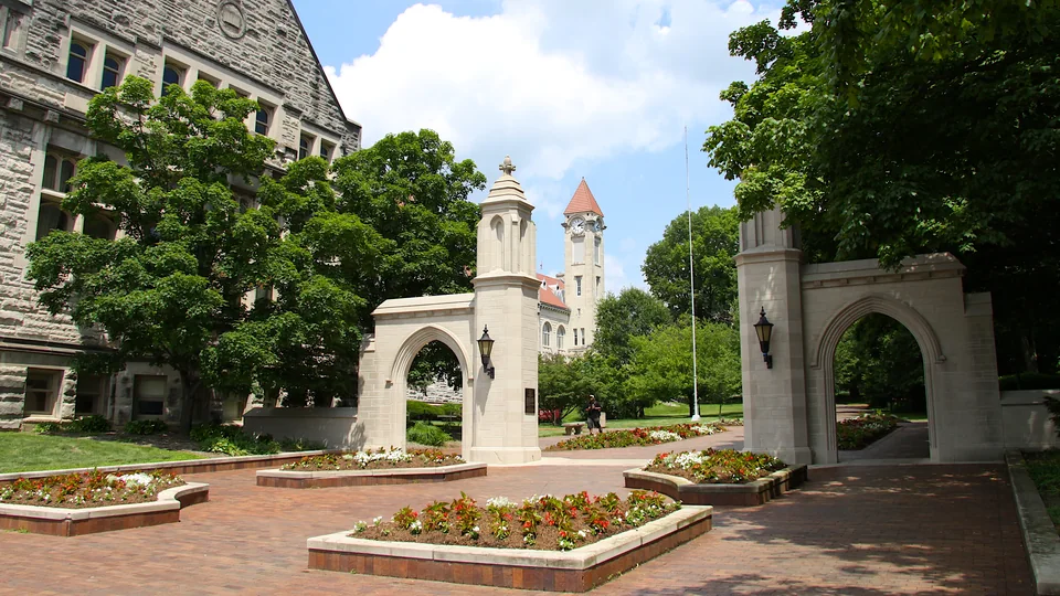 Two pale stone arches with lanterns, flowerbeds and a paved area in front, and a clock tower among trees in the distance