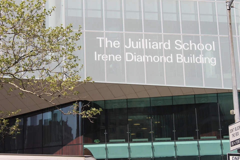Стеклянный фасад здания с надписью The Juilliard School над консольным выступом и ветви дерева слева