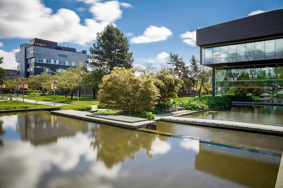 Стеклянное здание библиотеки Langara College у отражающего бассейна, на заднем плане — корпус с надписью Langara и деревья кампуса