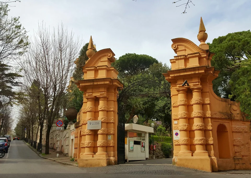 Оранжевые каменные столбы ворот с кованой решёткой и табличкой LUISS, слева улица с деревьями