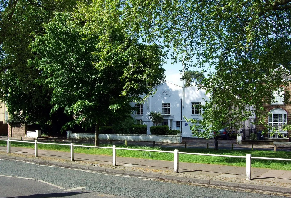 Белое двухэтажное здание с вывеской Ealing Studios за деревьями, перед ним газон и белое ограждение вдоль дороги
