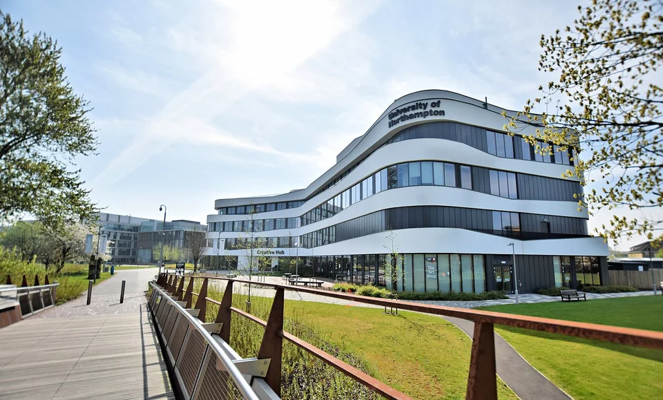 Волнообразное белое здание Creative Hub с надписью University of Northampton, газоны и пешеходный мост кампуса Waterside в солнечный день