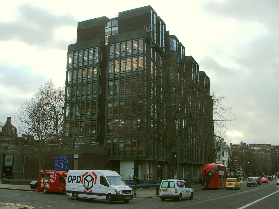 Тёмное многоэтажное здание Darwin Building Королевского колледжа искусств с ленточными окнами, перед ним — улица с фургонами и красным автобусом