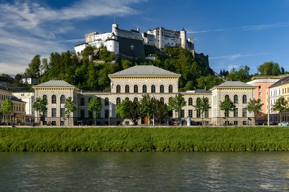 Классическое здание с надписью Universität Salzburg у реки, над ним на холме белая крепость