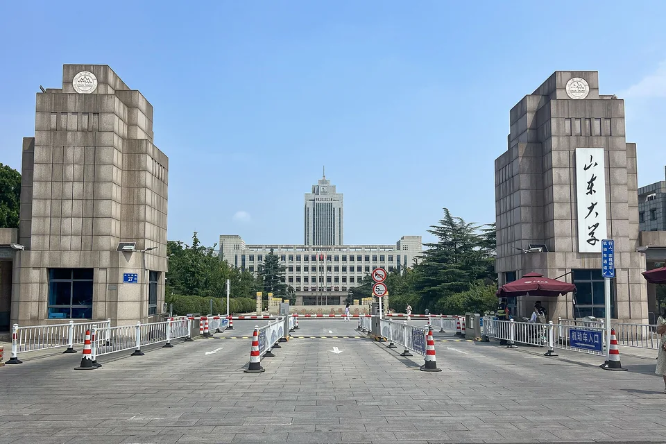 Две каменные башни ворот с вертикальной надписью «山东大学», за площадью с ограждениями и конусами видно высокое здание с часами