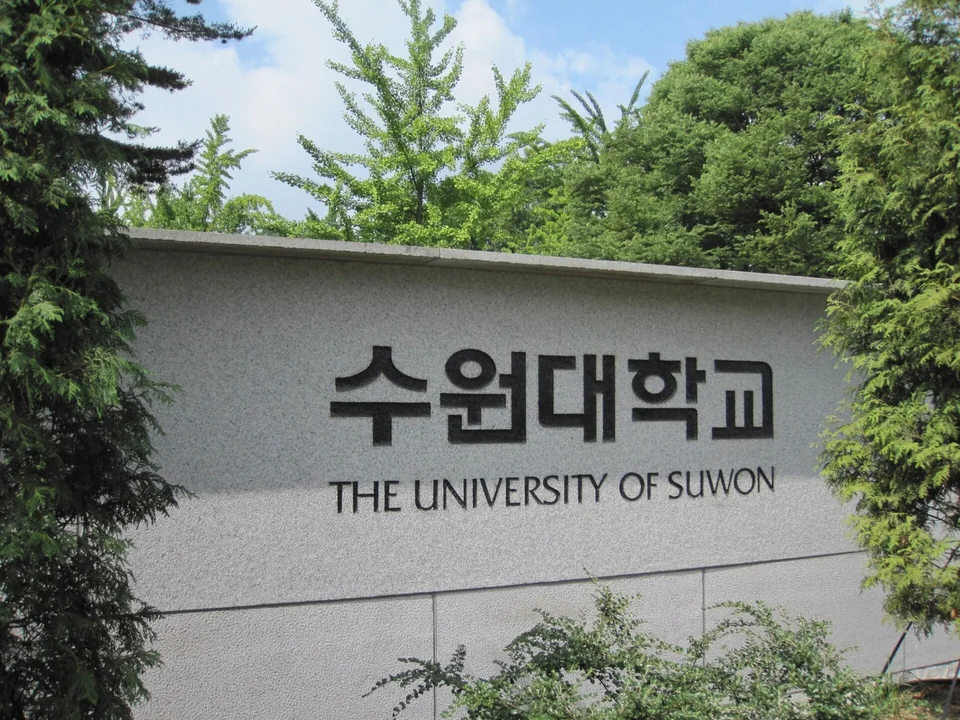 Серая гранитная стена с надписью на корейском и английском The University of Suwon в окружении зелёных деревьев